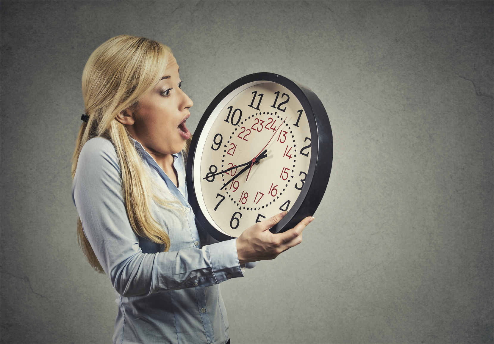 Closeup portrait woman, worker, holding clock looking anxiously, pressured by lack, running out of time, isolated on gray wall background. Human face expression, emotion, reaction